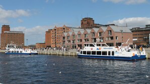 A waterfront scene with two boats docked at a pier, a large brick building with multiple windows, and a clear blue sky. - 3) Holiday apartment 4-room, upper floor, 110m² - Landhaus Alte Schule near (Alt Bukow)