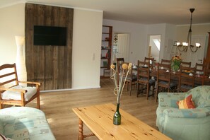 A living room with a wooden table, a sofa, a TV mounted on a wooden wall, and a dining area with chairs and a table.