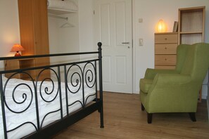 A bedroom with a bed, a green armchair, a wooden dresser, and a lamp.
