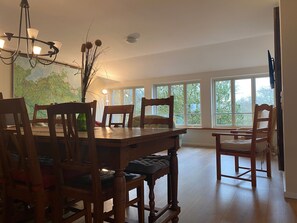 A dining room with a wooden table and chairs, a chandelier, and large windows with a view of trees.