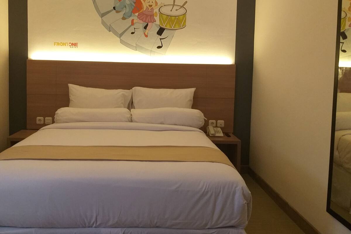 Una habitación de hotel con una cama grande, ropa de cama blanca, un cabecero con un reloj, una mesita de noche con un teléfono y un mural en la pared.