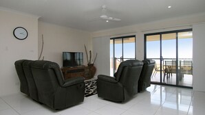 A living room with two reclining chairs, a TV, and a wooden cabinet. There is a large window with a view of the beach. - 17 Sea Esplanade, Elliott Heads (ELLIOTT HEADS)