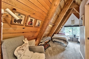 An attic bedroom with wooden walls, a sloped ceiling, and a large window. There are two beds, a sofa, and a deer sculpture.