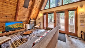 A cozy cabin interior with a large TV, wooden walls, a sofa, and a view of the forest through large windows. - Külhaus Cabin - Romantic A-frame Adventure, BBQ (South Lake Tahoe)