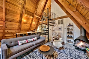 Una habitación estilo loft acogedora con techo de madera, un sofá gris, una escalera de caracol y un hogar a leña.