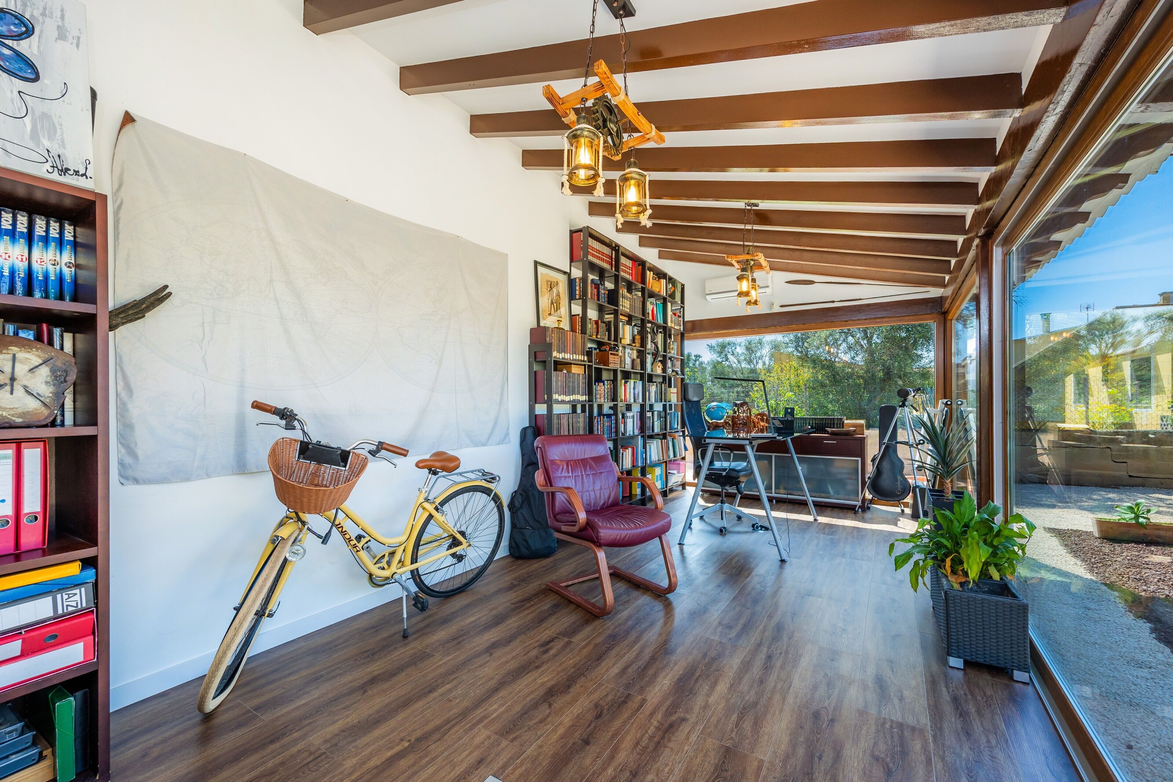 Ein Zimmer mit Holzbalken, einem Bücherregal, einem Fahrrad, einem Schaukelstuhl und Blick ins Freie.