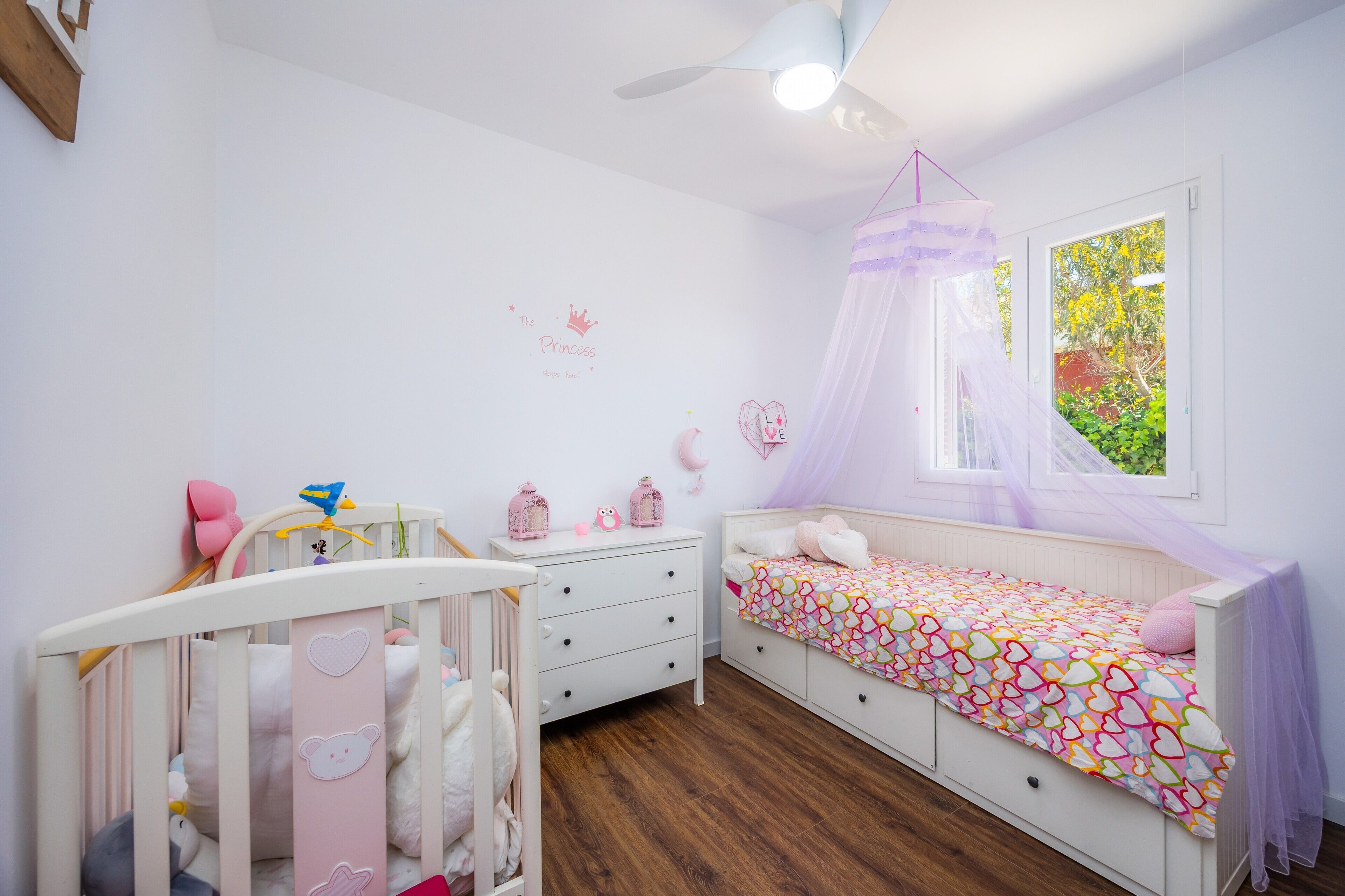 A child's bedroom with a crib, a bed with a canopy, and a window with a view of greenery.