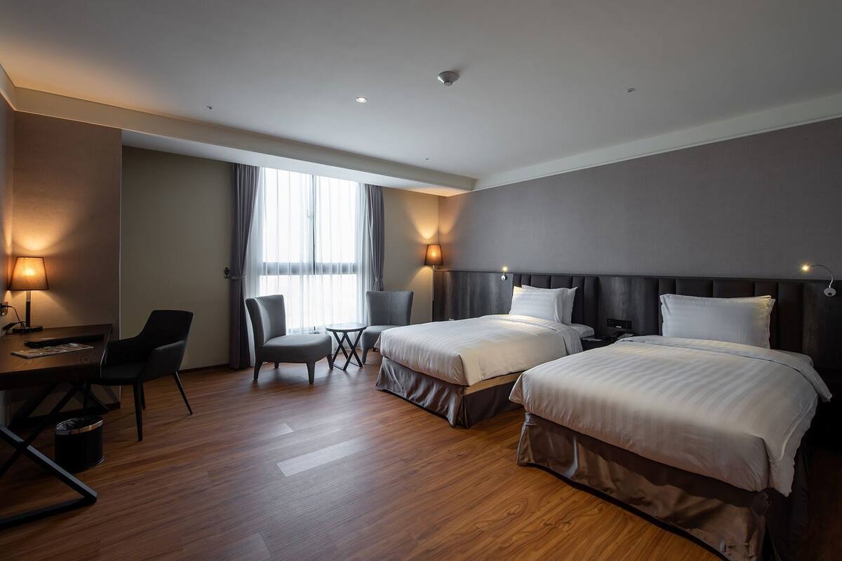 Camera d'albergo con due letti, una scrivania, una sedia, un tavolino e una finestra con tende.