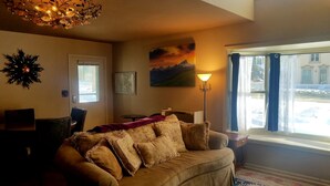A cozy living room with a brown sofa, a painting of a mountain landscape, and a window with curtains. - Gowdy Apartment. Fenced yard pet friendly , modern, clean. Gowdy Properties (Lake City)
