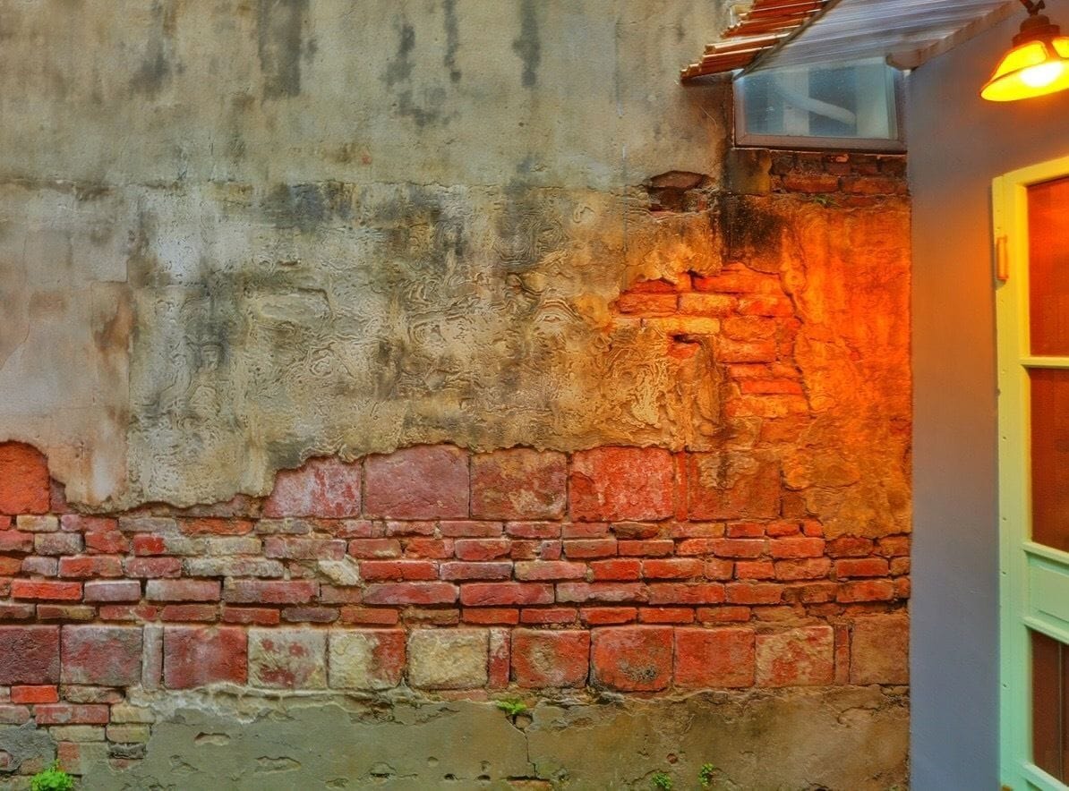 A weathered brick wall with a rough texture and a partially visible window.