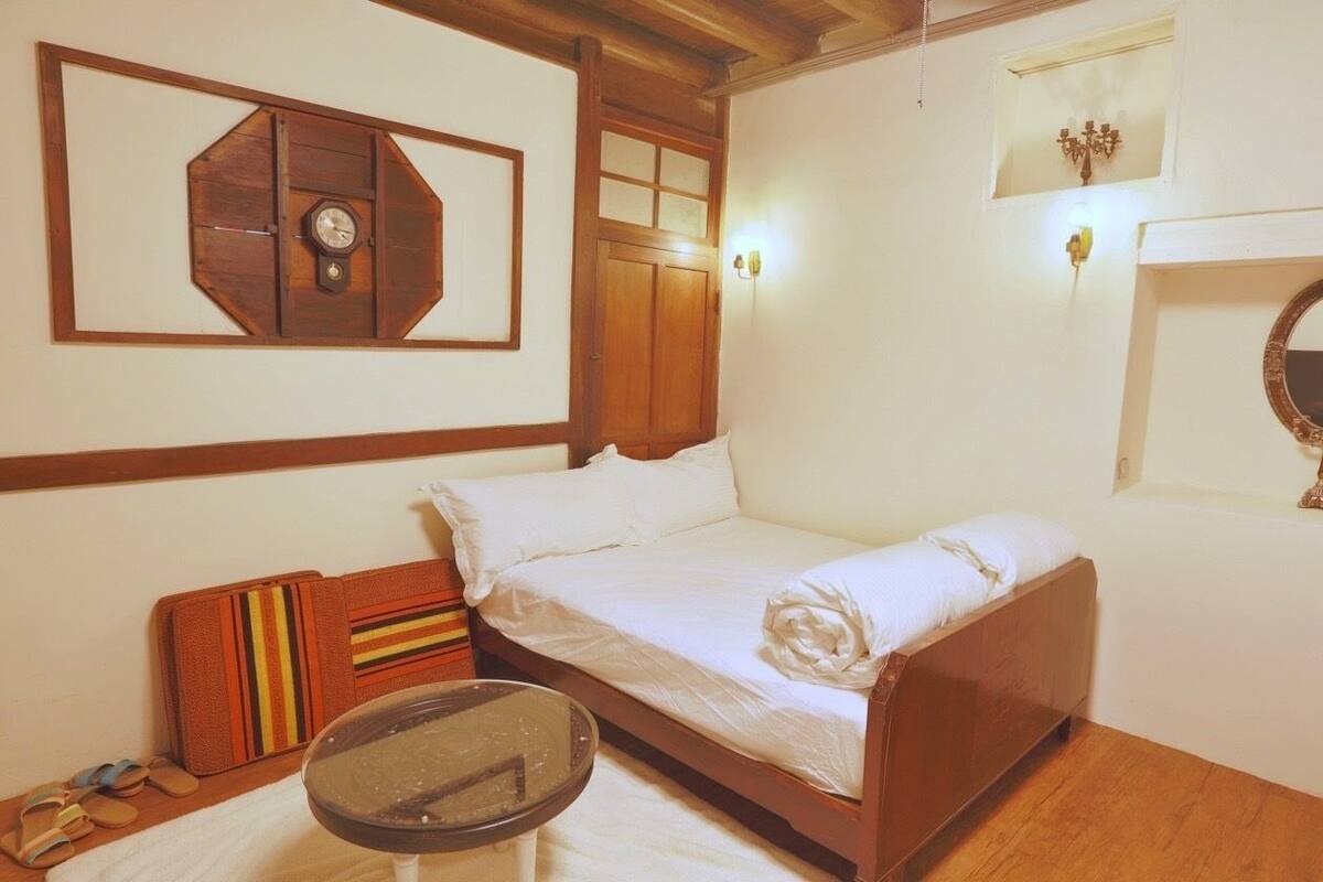 A single bed with a wooden headboard, a bedside table, a wall-mounted clock, and a small mirror.
