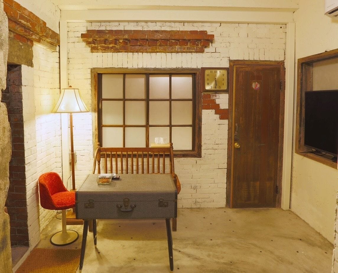 A small room with a brick wall, a wooden door, a window with blinds, a table with a suitcase, a red chair, and a lamp.