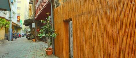 A narrow street with buildings on either side, a bamboo fence, and a potted plant.