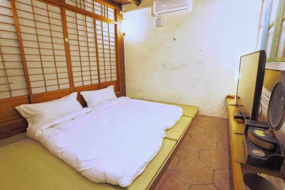 A traditional Japanese-style room with tatami flooring, a low wooden bed with white bedding, a shoji screen, and a small kitchen area with a stove.