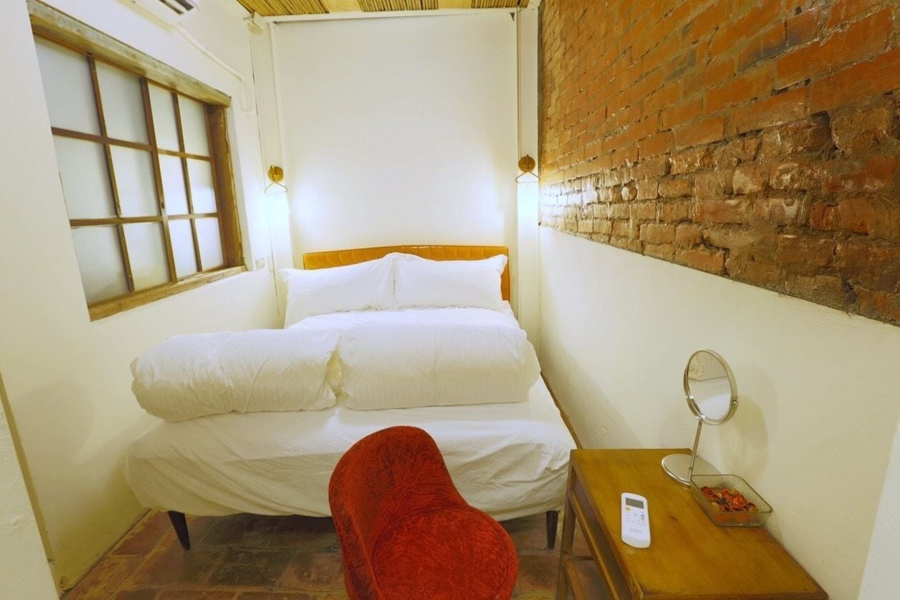Une petite pièce chaleureuse, avec un lit simple, une chaise rouge, une table de chevet en bois et un mur en briques.
