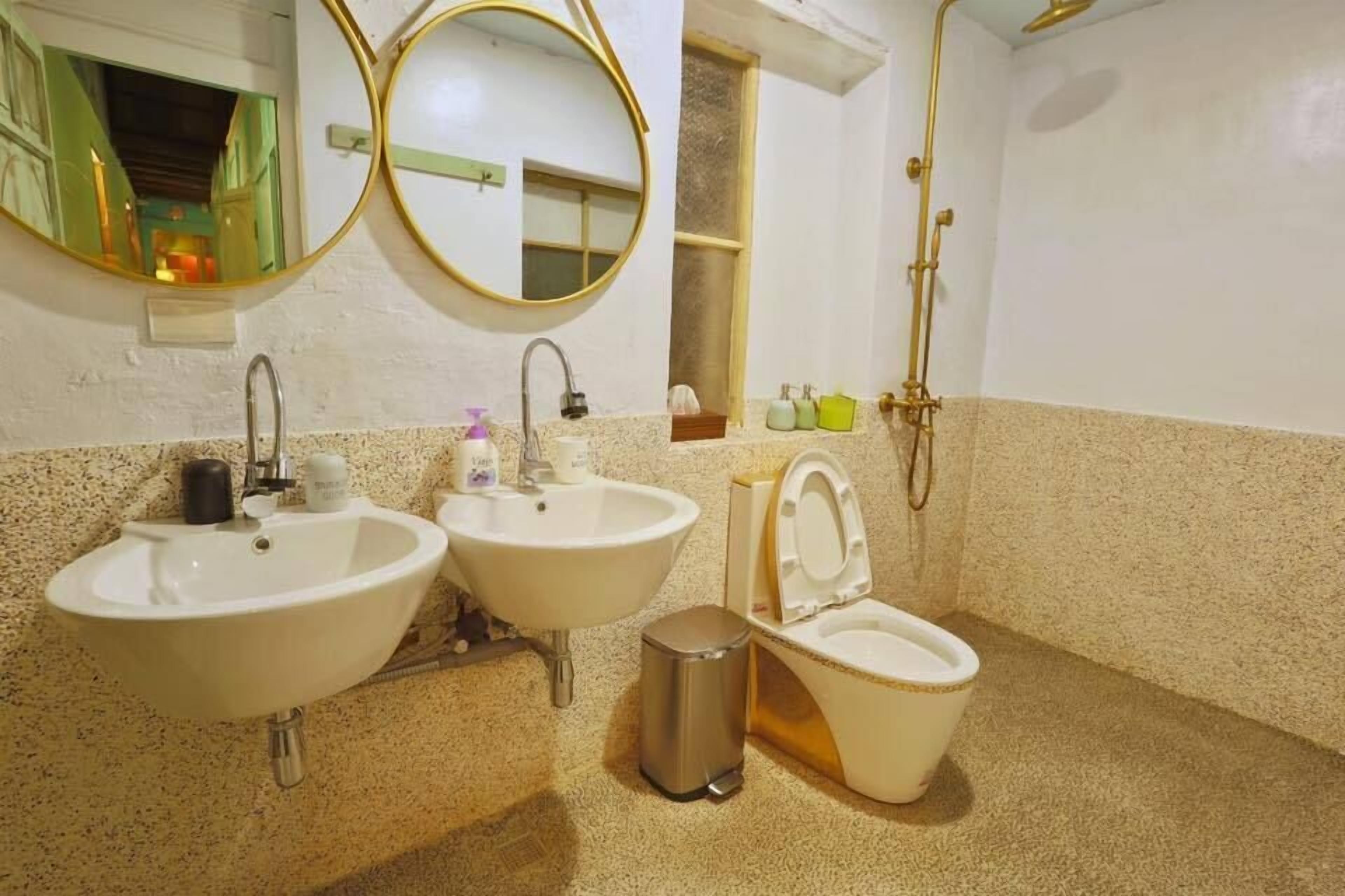 A bathroom with two sinks, a toilet, a trash bin, a mirror, and a shower.