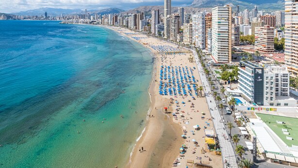 A coastal cityscape with a beach lined with blue umbrellas and a high-rise hotel. - Don Miguel 1 18-A (Benidorm)
