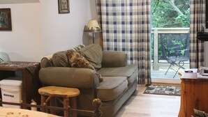 Kodikas olohuone, jossa on sohva, pieni puinen pöytä ja lamppu. Ikkunasta, jossa on verhot, avautuu näkymä ulos.
