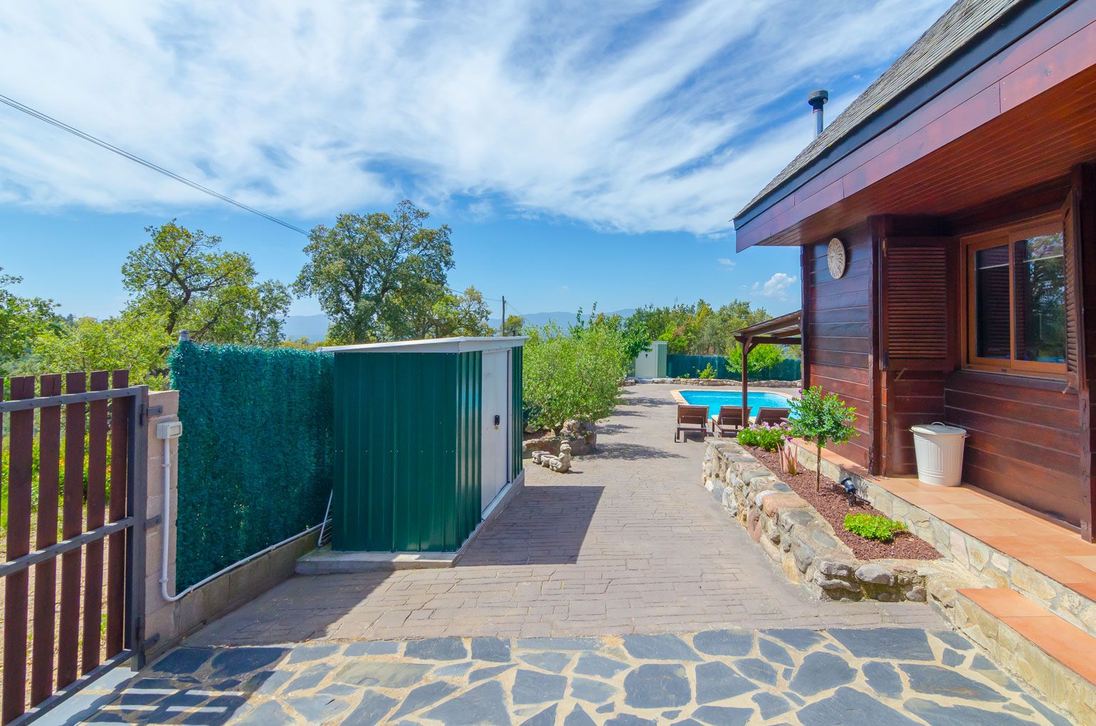 A pathway leading to a wooden building with a pool, stone paving, and a green storage shed.