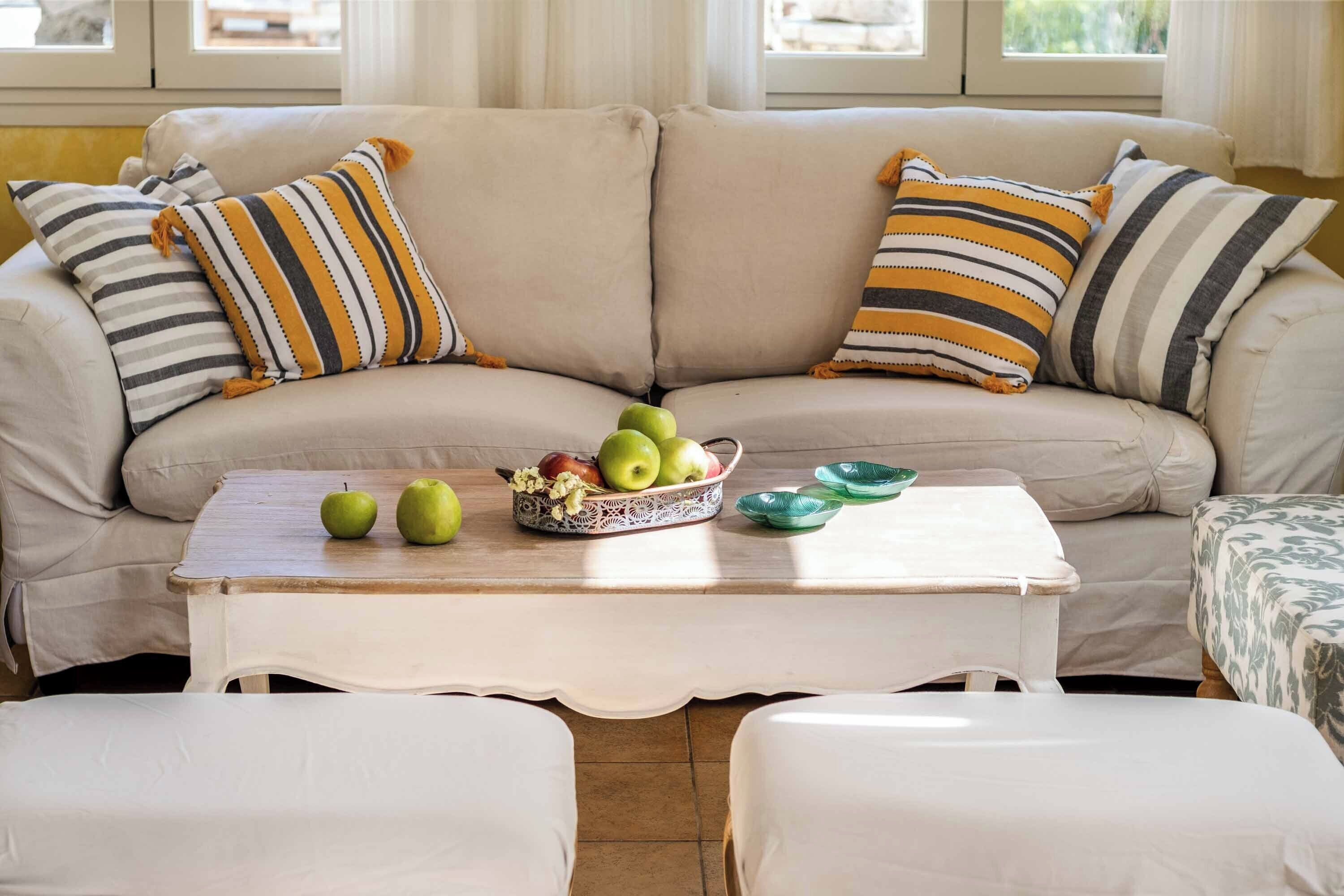 A beige sofa with striped pillows, a white table with apples and a bowl of fruit, and a patterned throw on a green chair.
