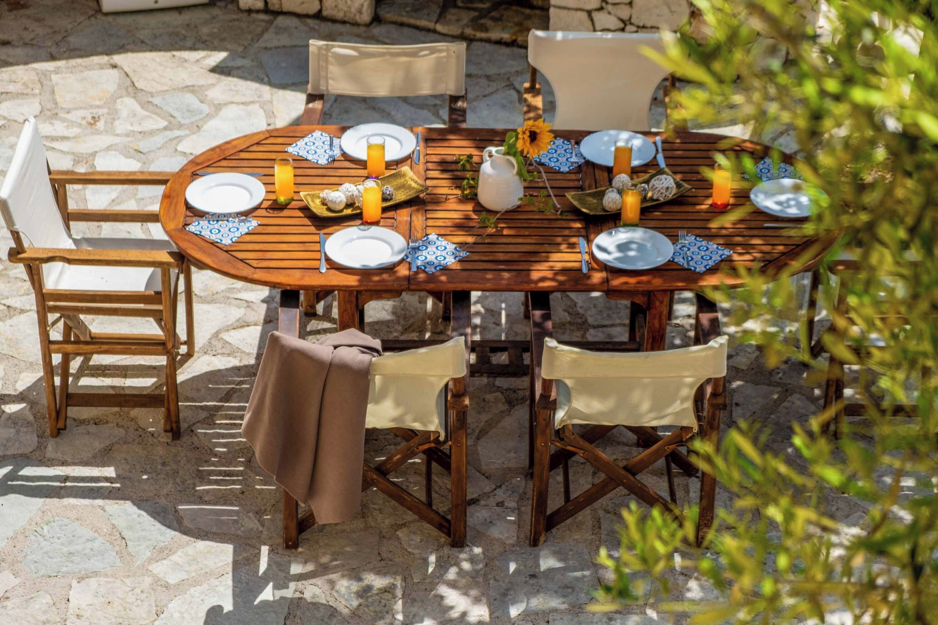 Mesa de madera al aire libre, preparada para cuatro personas, con platos, cubiertos y bebidas, rodeada de sillas y un piso de piedra.