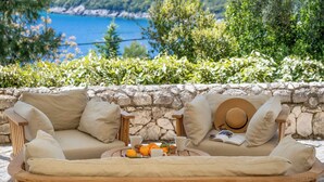 A wooden sofa with cushions, a stone wall, and a view of the sea. - Villa Elpida (Lefkada)