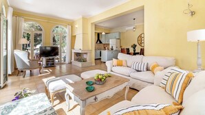 A spacious living room with a sofa, coffee table, and TV. A kitchen is visible in the background. - Villa Elpida (Lefkada)