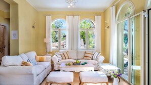 A cozy living room with a sofa set, a coffee table, and a dining table. There are large windows with curtains, a chandelier, and a view of greenery outside. - Villa Elpida (Lefkada)