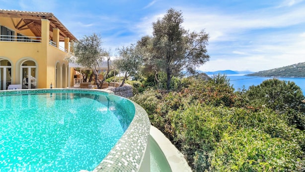 A poolside view of a Mediterranean-style villa with a clear blue pool, surrounded by lush greenery and overlooking a scenic coastline. - Villa Elpida (Lefkada)