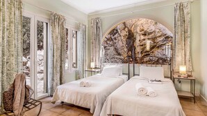 A hotel room with two beds, a large window with curtains, a view of a rocky landscape, and a wall mural. - Villa Elpida (Lefkada)
