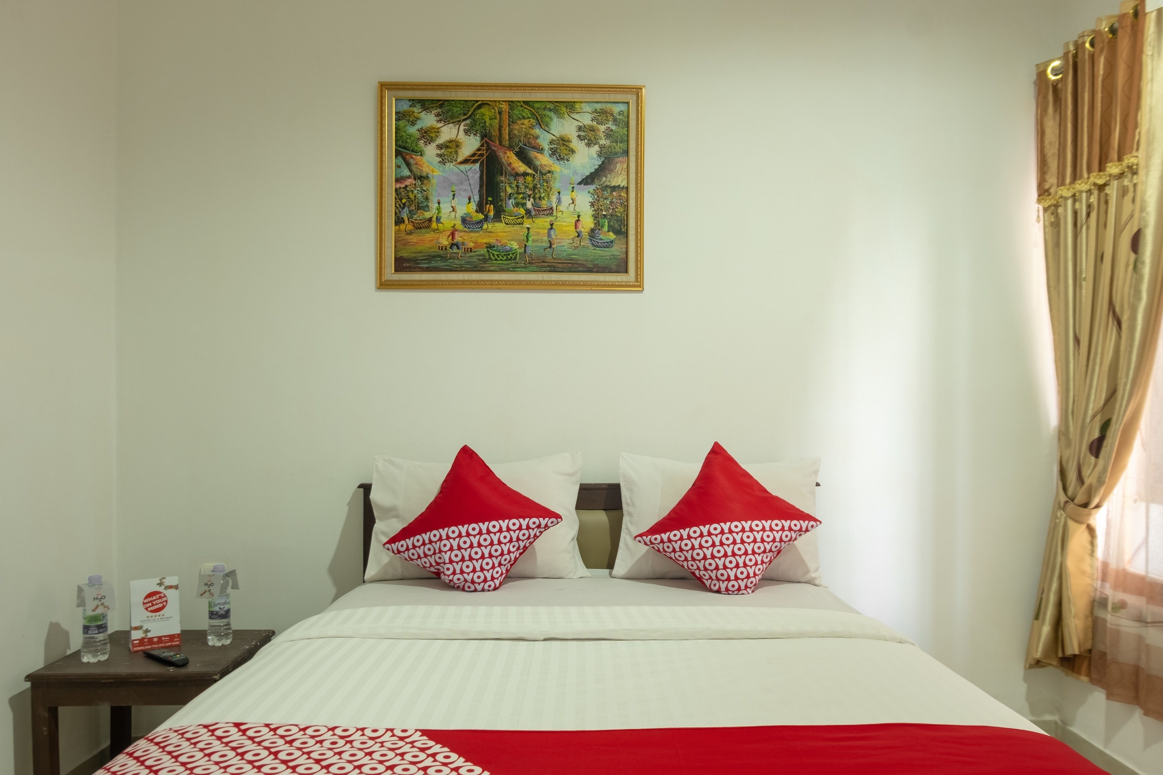 A hotel room with a bed, two red and white patterned pillows, a nightstand with a remote control and water bottles, and a painting on the wall.