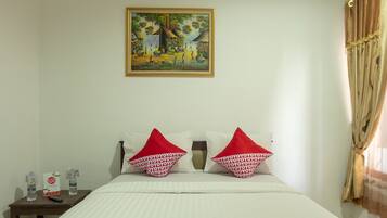 A hotel room with a bed, two red and white patterned pillows, a nightstand with a remote control and water bottles, and a painting on the wall.