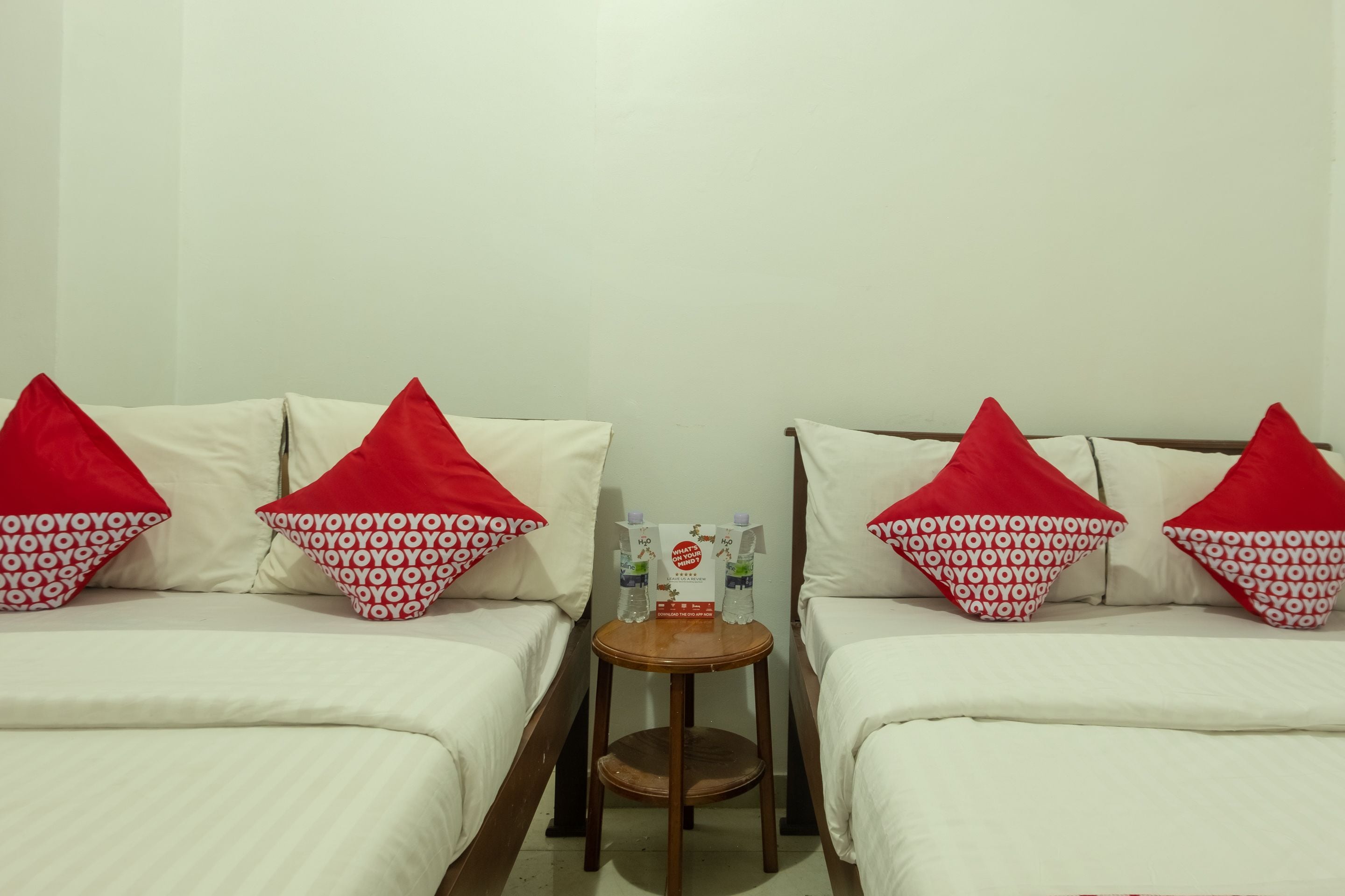 Habitación de hotel con dos camas, cada una con ropa blanca y almohadas rojas, una mesita de madera con una botella de agua y una tarjeta, y una pared blanca lisa.