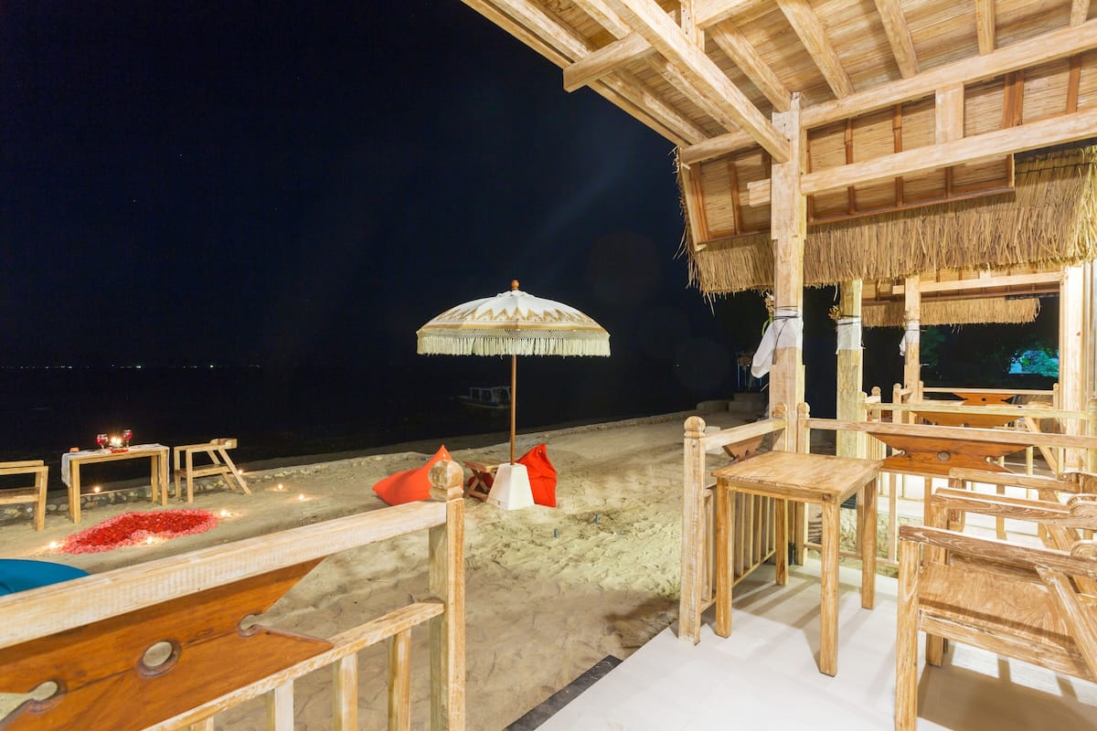 Un espacio de descanso frente al mar con mesas y sillas, una estructura de techo de paja y un paraguas decorativo.