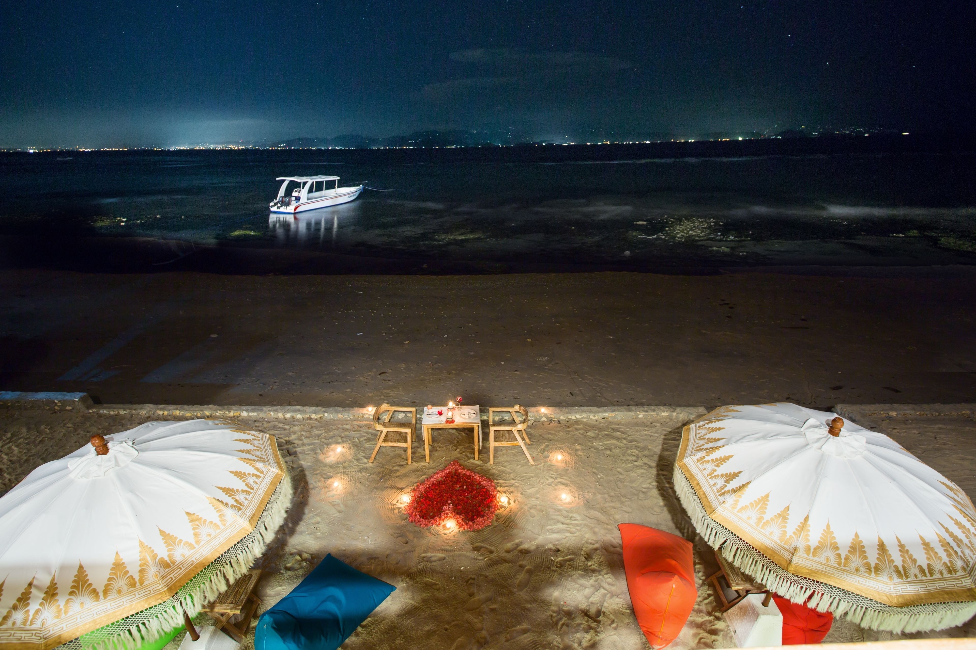 Un espacio junto a la playa con una mesa y sillas, un barco al fondo y sombrillas.