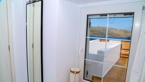 Ein Badezimmer mit einem großen Spiegel, einer kleinen Mülltonne und einer Glastür, die auf einen Balkon mit Blick auf das Meer und die Hügel führt.