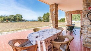 An outdoor patio with a table set for dining, stone pillars, wicker chairs, and a view of a grassy area with trees and a clear sky. - Lovely home in Sassetta with swimming pool (Sassetta)