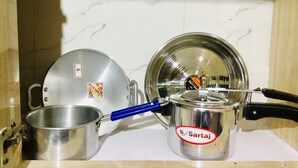 Une cuisine avec des casseroles et des poêles en acier inoxydable, une poêle à manche bleu et une casserole portant l’inscription « Sartaj ».