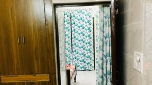 Une pièce avec une armoire en bois, un lit recouvert d’une couvre-lit à motifs floraux, un rideau à motifs et un sol carrelé.