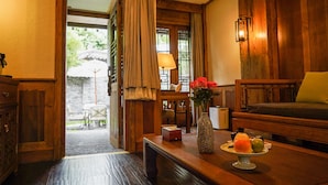 A wooden interior with a sofa, a table with a vase and fruit, and a view of an outdoor area with a chair and umbrella. - Songtsam Linka Lijiang (Lijiang)