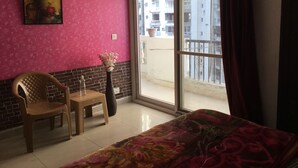 A room with a pink wall, a large window with a view of buildings, a chair, a small table, and a vase with flowers. - HomeNow - Full Kadambari - Botanical Garden Metro (Noida-)