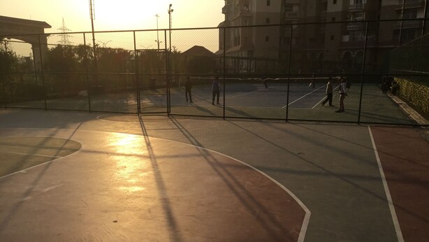 An outdoor basketball court with a fence, surrounded by buildings. - HomeNow - Full Kadambari - Botanical Garden Metro (Noida-)