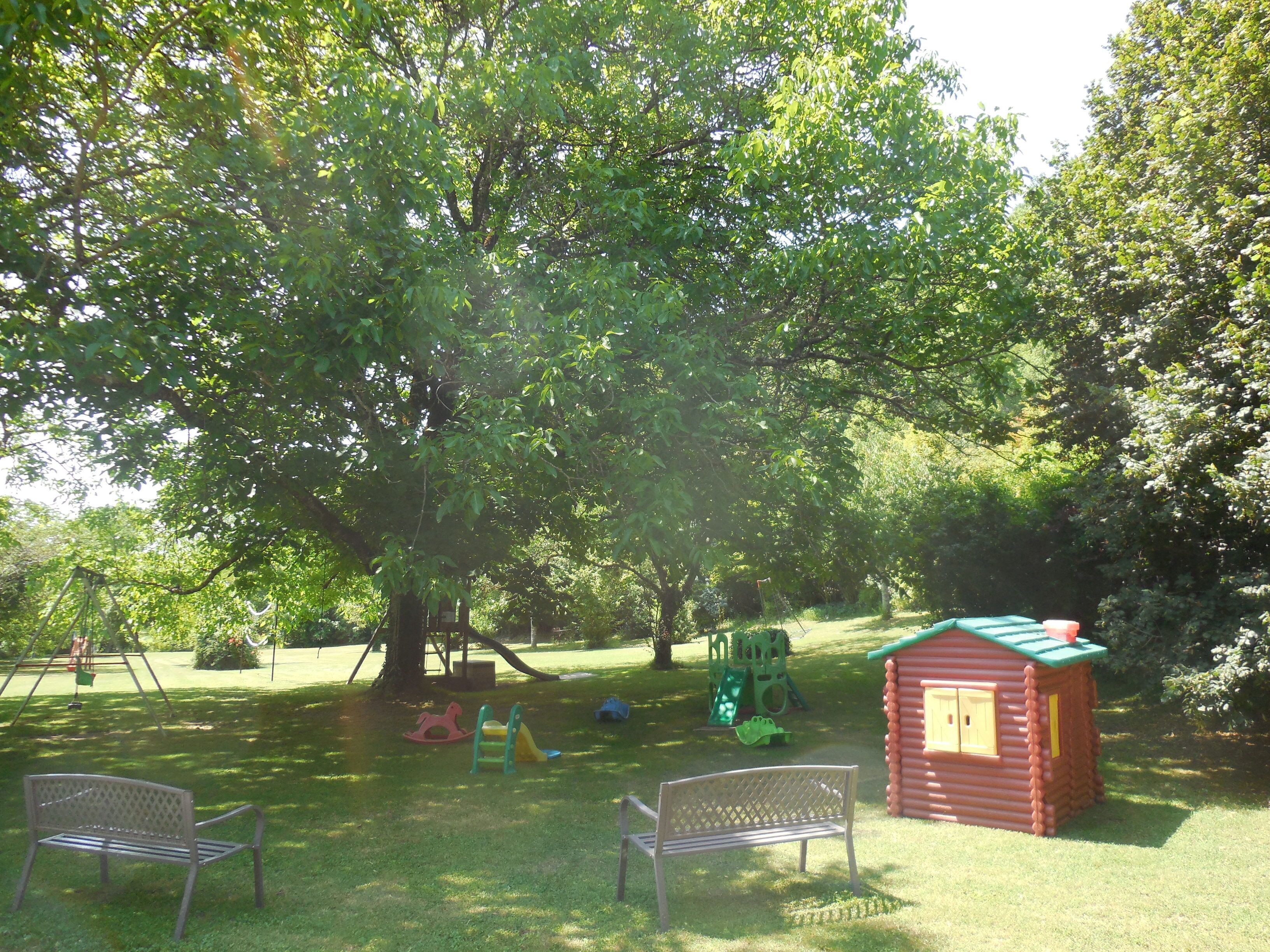 A park with a swing set, a slide, a small playhouse, and two benches.