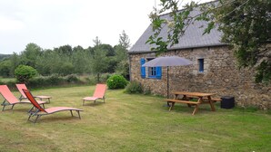 Un edificio de piedra con techo inclinado, una mesa de picnic y dos sillones de jardín en un espacio al aire libre cubierto de césped.