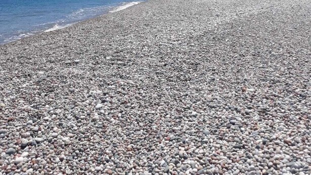 Pikkukivinen ranta, jonka yllä on kirkas sininen taivas ja kauempana näkyy kukkuloita.