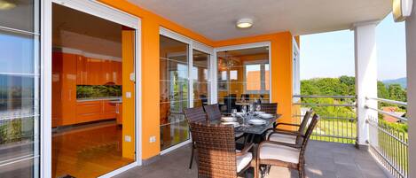 A balcony with a dining table and chairs, a view of the outdoors, and a kitchen area with orange cabinets.