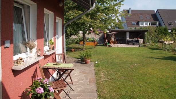 Un edificio rosso con un patio attrezzato con tavolo e sedie, immerso in un prato curato e ombrado da un albero.