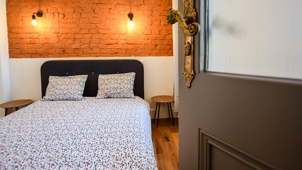 A bedroom with a brick wall, two wall-mounted lights, a bed with a floral-patterned bedspread, a wooden nightstand, and a small round table. - MB2- Gwarna 11 (Wroclaw)
