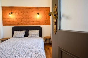 Un dormitorio con pared de ladrillo, dos luces fijadas a la pared, una cama con colcha estampada con motivos florales, una mesita de noche de madera y una pequeña mesa redonda.