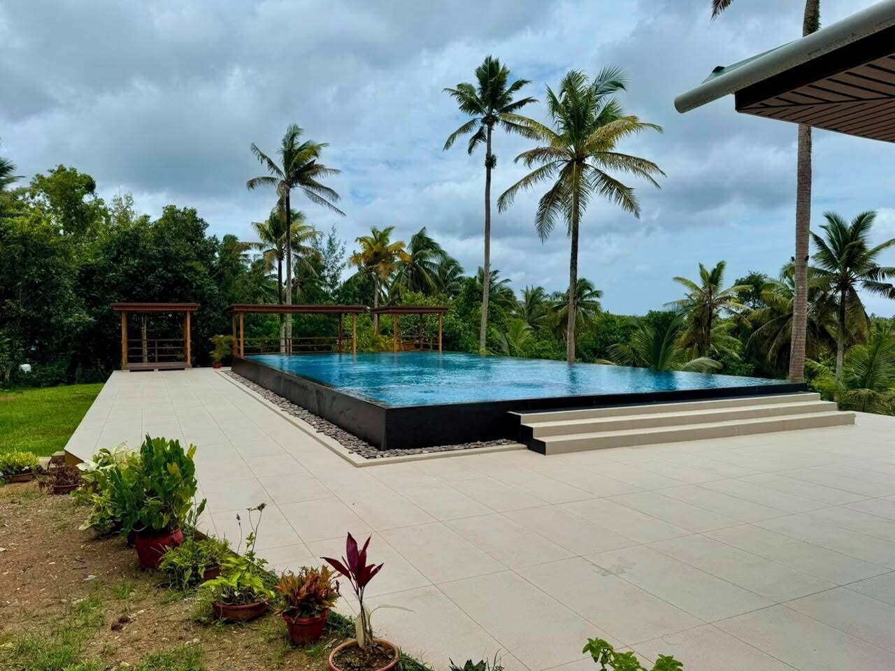 Uma piscina com deck ladrilhado, cercada por palmeiras e um céu nublado.